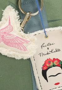 Sleutelhanger met een roze gevleugeld vogels ontwerp op stof en een label met een door Frida Kahlo geïnspireerde illustratie, tegen een groene textiel achtergrond.