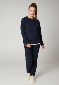 Felpa blu navy e pantaloni coordinati in tessuto morbido. Presenta un collo rotondo e polsini a coste. Indossata con sneakers beige.