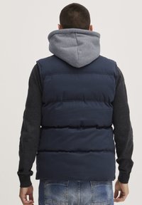 Navy puffer vest with adjustable gray hood, padded design, and textured gray sleeves. Casual denim jeans complete the outfit.