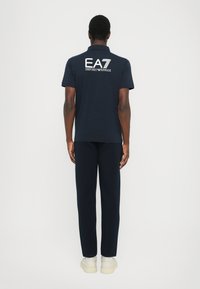 Pólo azul marinho com o logo EA7 nas costas, combinado com calças azul marinho e sapatos de cor clara. Textura suave, ajuste padrão.