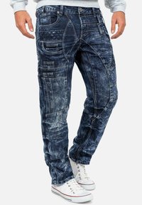 Pantaloni cargo in denim blu lavato con multiple tasche, vari pattern di cucitura e una vestibilità affusolata, abbinati a sneakers bianche.