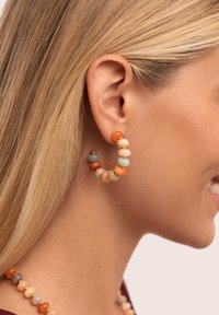 Boucles d'oreilles en perles en forme d'anneau, arborant des couleurs variées, notamment orange, vert et crème. Le design comprend des accents dorés et une forme circulaire.