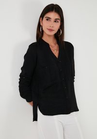 Black long-sleeve button-up shirt with a v-neck, featuring two front pockets and a textured finish. Styled with white pants.