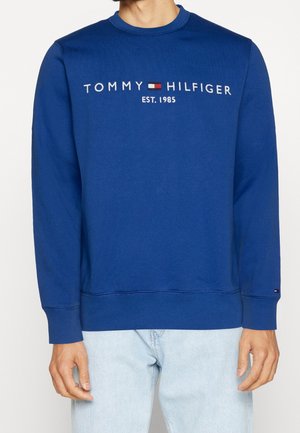 Mężczyzna noszący niebieską bluzę Tommy Hilfiger z logo i napisem „EST. 1985”, w połączeniu z jasnoniebieskimi dżinsami, na jednolitym tle.