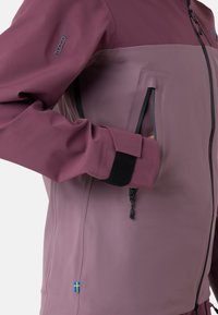 Bordeauxrød softshell-jakke med glat tekstur, der har en lynlåslomme, justerbar håndledsmanchet og et lille svensk flag-detalje.