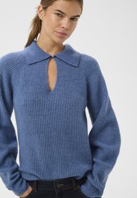 Modra pletena pulover s ovratnikom in detajlom v obliki ključavnice. Ima rebrasto teksturo in sproščene rokave. V kombinaciji s temnimi jeans hlačami.