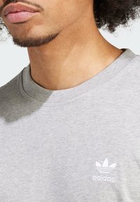 Šedé bavlněné tričko s žebrovaným kulatým výstřihem a vyšitým bílým logem Adidas trefoil na levé hrudi. Měkký, ležérní design.