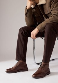 Hnedé kožené členkové topánky s hladkým povrchom, zaoblenou špičkou a jemnými švami. Kombinované s tmavohnedými nohavicami a textúrovanou košeľovou bundou.