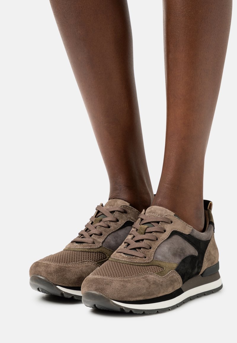 Baskets en daim marron et noir avec des détails en mesh portées sur des pieds à peau foncée, présentées sur un fond clair uni.