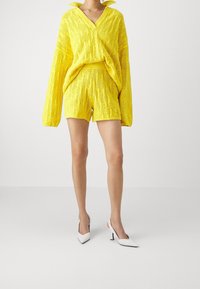 Conjunto de dos piezas de punto amarillo que incluye un top holgado de manga larga y pantalones cortos, ambos con patrones texturizados. Combinado con zapatos de tacón blancos.