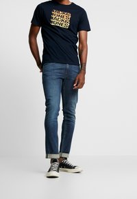 Muž v námořnickém tričku Jack & Jones, s manžetovými modrými džínami a černými nízkými plátěnými teniskami na bílém pozadí.