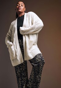 Flauschige, cremefarbene Strickjacke mit Kabelmustern, ausgestattet mit zwei Taschen. Kombiniert mit lockeren Leopardenmusterhosen in Schwarz- und Beigetönen.