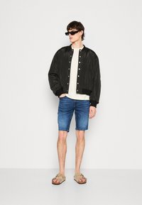 Giubbotto bomber nero con bottoni, camicia bianca, pantaloni shorts in denim blu e sandali beige. Modella che indossa occhiali da sole neri, in piedi contro uno sfondo neutro.