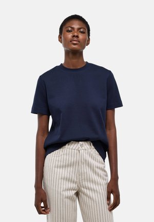 Femme portant un t-shirt bleu marine à manches courtes et un pantalon beige taille haute avec de fines rayures noires verticales sur fond blanc.