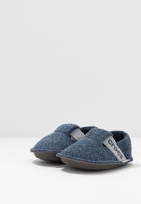 Chaussons en laine bleu doux à enfiler avec des semelles grises et une bande grise étiquetée "crocs", placés côte à côte sur une surface blanche.