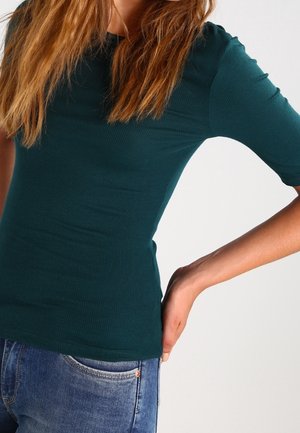 Person iført en tætsiddende mørkegrøn top med trekvartærmer og blå jeans, poserer med den ene hånd hvilende på hoften.