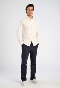 Camicia bianca in avorio con maniche lunghe, abbinata a pantaloni blu navy. Il modello indossa sneakers bianche, in piedi con le mani nelle tasche.