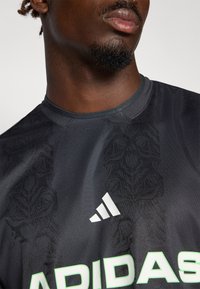 Zwarte sporttrui met een subtiele gemusterde textuur, ronde halslijn, wit Adidas-logo en groene merkletters op de voorkant.