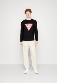 Zwarte longsleeve T-shirt met een rood driehoekig logo waarop "GUESS" en een vraagteken staat, gecombineerd met crèmekleurige broeken en witte sneakers.