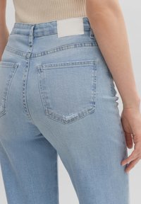 Detalle de unos vaqueros de denim azul claro con bolsillos traseros que lleva puesta una persona con una blusa de punto beige, mostrando costuras y un ligero desgaste en los bolsillos.