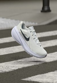 Ανοιχτό γκρι Nike αθλητικό παπούτσι με υφή στο πάνω μέρος, μαύρη λωρίδα τόνωσης και άνετη λευκή σόλα, τοποθετημένο πάνω από διάβαση πεζών.