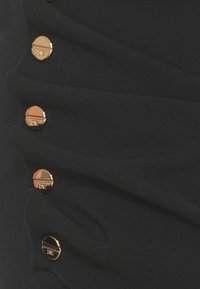 Drappe di tessuto nero con texture arricciata; sei bottoni metallici dorati disposti verticalmente, ognuno con un design circolare piatto.