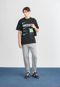 T-shirt preta de manga curta com texto gráfico verde e branco, calças de moletom cinza com o logo laranja da Nike, e sapatilhas de desporto pretas.