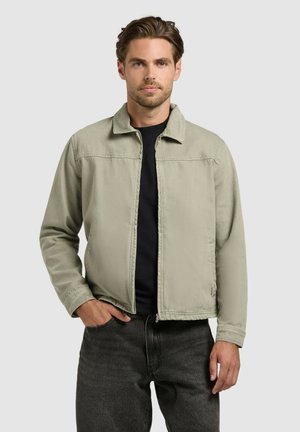 Mees helerohelise lukuga jopes musta särgi kohal, tumeda halli teksadega, seisab ühe käega taskus tavalise tausta ees.