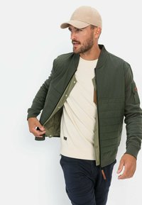 Olijfgroene bomberjack met gevoerde secties, drukknoopsluitingen en een binnenvoering. Draagt over een lichte trui en gecombineerd met donkere broeken. Beige pet.