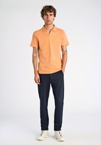 Polo shirt arancione a maniche corte con colletto, abbinato a pantaloni blu e sneakers bianche. L'outfit presenta un design semplice e pulito.