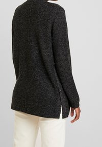 Dlouhý černý svetr s texturovaným pletením, s vysokým límcem, postranními rozparky a volným střihem. Kombinováno s off-white kalhotami.