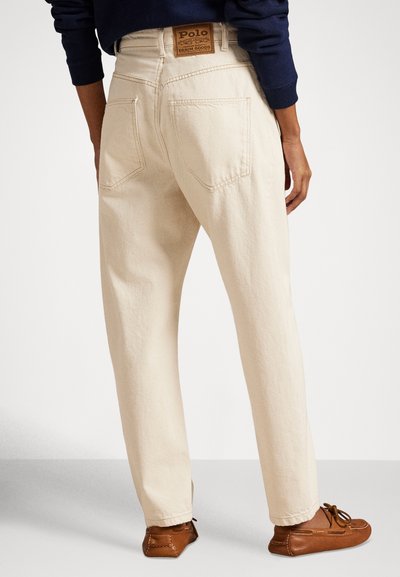 Μπεζ τζιν παντελόνια με ίσια γραμμή στο πόδι, πίσω τσέπες και δερμάτινη ετικέτα με λογότυπο στη μέση. Φορεμένα με καφέ μοκασίνια.