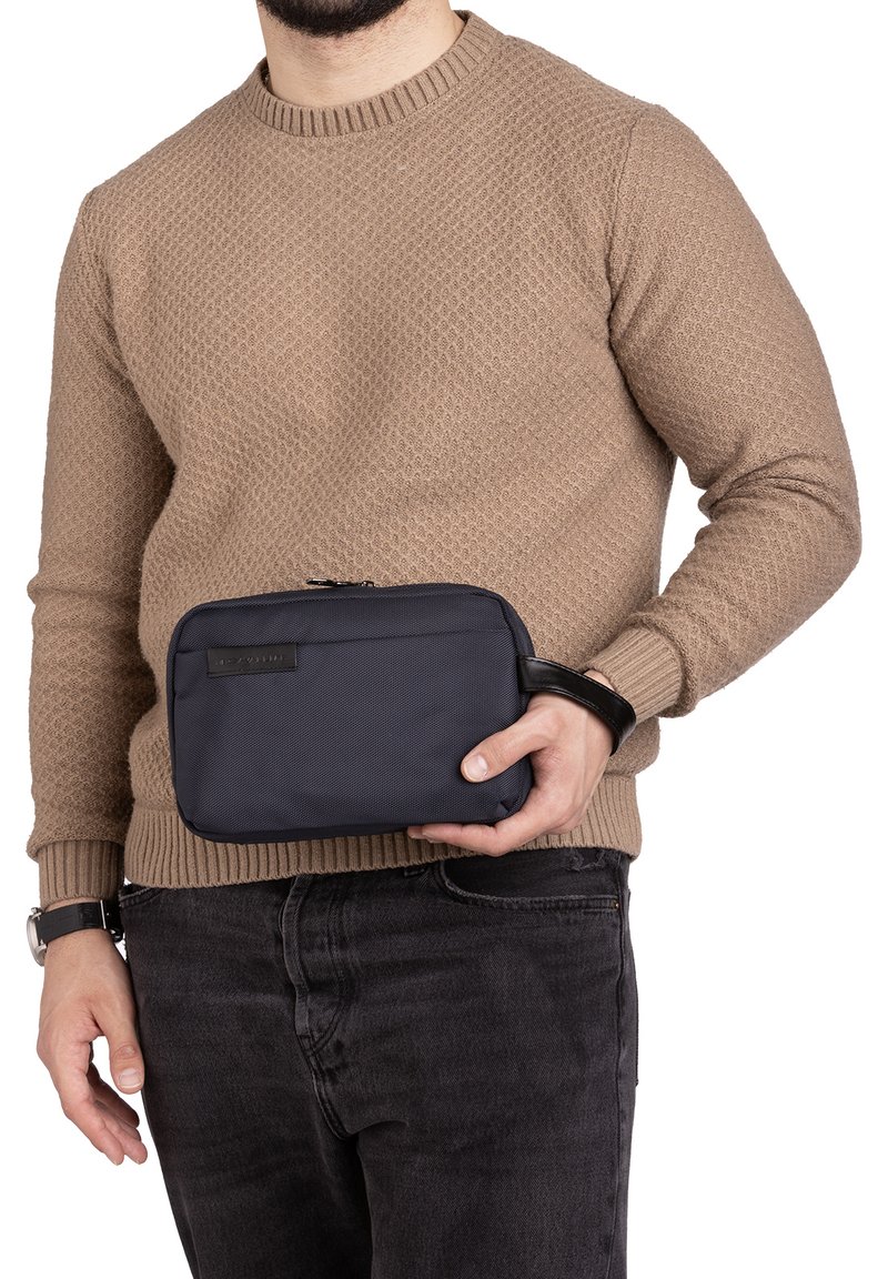 Borsa da toeletta in tessuto di colore blu navy con chiusura a cerniera e una tracolla nera, tenuta da una persona con un maglione di lana color beige.