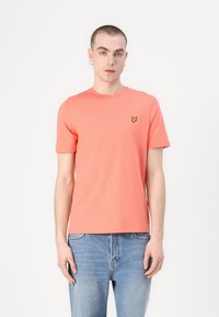 Korallfärgad kortärmad T-shirt i bomull, med rund halsringning och en liten gul fjärilslogotyp på vänster bröst.
