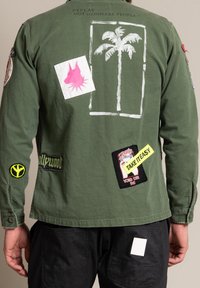 Olivgrön långärmad skjorta med olika lappar, inklusive en palmträd design, ljusrosa detaljer och broderad text.