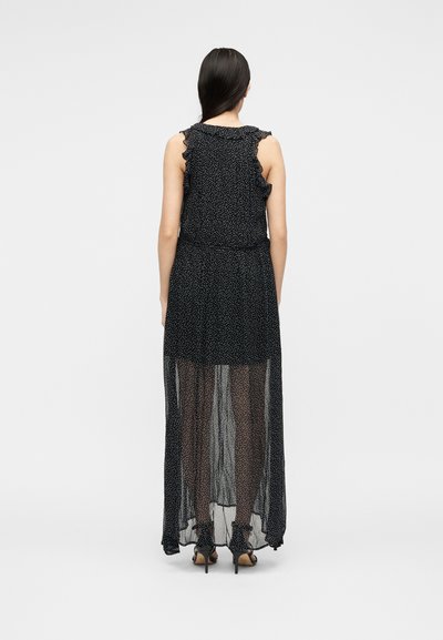 Γυναίκα με μακριά σκούρα μαλλιά που φοράει αμάνικο μαύρο φόρεμα με λευκά πουά και διαφανές μακρύ φόρεμα, στέκεται με την πλάτη στην κάμερα.