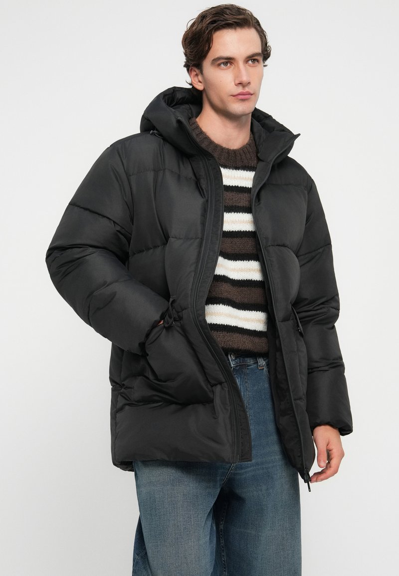 Schwarz gepolsterte Winterjacke mit Kapuze, mit einem Reißverschluss und Taschen an den Seiten, getragen über einem gestreiften Pullover und blauen Jeans.