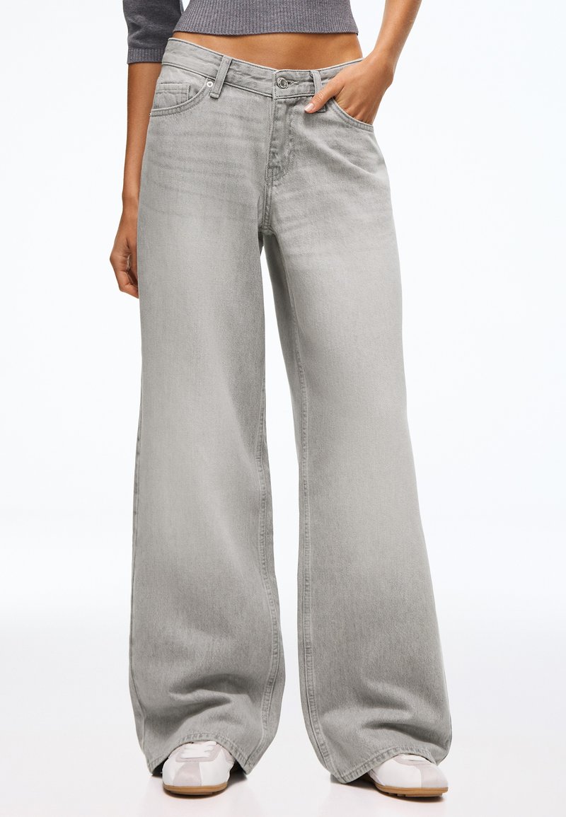 Personne portant un jean gris clair taille haute à jambes larges avec poches avant, associé à des baskets blanches et un pull gris court.