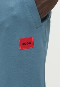 Világoskék pamut melegítőnadrágok piros téglalap alakú címkével, amelyen fekete "HUGO" felirat olvasható. Az anyag sima textúrájú, kényelmes, lezser szabású.