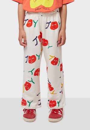 Un pantalon de survêtement blanc avec un motif répétitif de cerises rouges souriantes et de lettres colorées, associé à des baskets rouges.
