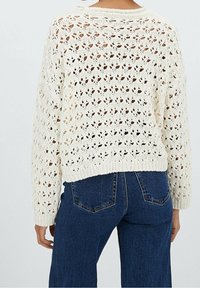 Pletený svetr v barvě slonové kosti s volným, otevřeným vzorem; má zaoblený výstřih a dlouhé rukávy, ve spojení s modrými džíny z denimu.