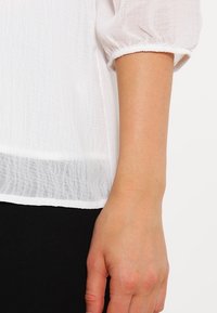Blusa bianca strutturata con maniche arricciate, caratterizzata da un sottile dettaglio sull'orlo. Mano su uno sfondo nero per il contrasto.