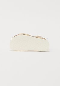 Platt sandal med en texturerad vit gummisula. Har två justerbara remmar i guldton över en brun bas, vilket ger en minimalistisk design.