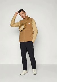 Tan fleece vest met ritszakken, gedragen over een beige langemouwen shirt, gecombineerd met zwarte broek en witte sneakers; accessoires zijn onder andere een zonnebril.