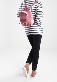 Rosa Samt-Rucksack mit metallischen Nieten, runder Form, verstellbaren Trägern und einer vorderen Reißverschlusstasche. Getragen mit einem gestreiften Oberteil und schwarzen Hosen.