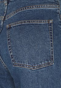 Jeans i mörkblå denim med en synlig bakficka. Har orange sömmar, en jämn textur och ett lätt blekt mönster.