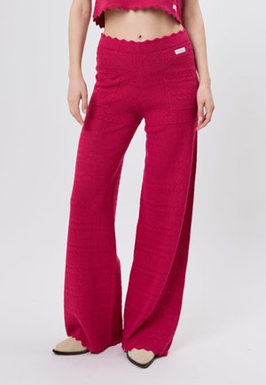 Frau trägt hellrosa, weit geschnittene, strukturierte Strickhose mit Fronttaschen und wellenförmigen Kanten, kombiniert mit beigen spitzen Schuhen.