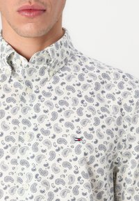 Bela srajca z gumbi in mornarsko modrim paisley vzorcem, ki ima majhen rdeč in moder logotip na levem žepu na prsih. Gladko bombažno blago.