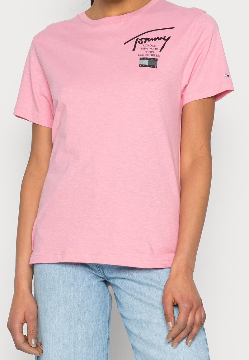 Jasnoróżowa bawełniana koszulka, krótki rękaw, okrągły dekolt, z czarnym nadrukiem logo na przodzie. Klasyczny krój o gładkiej teksturze.