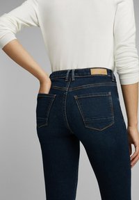 Personne portant un jean taille haute bleu foncé et un haut blanc à manches longues, main placée dans la poche arrière, fond gris neutre.